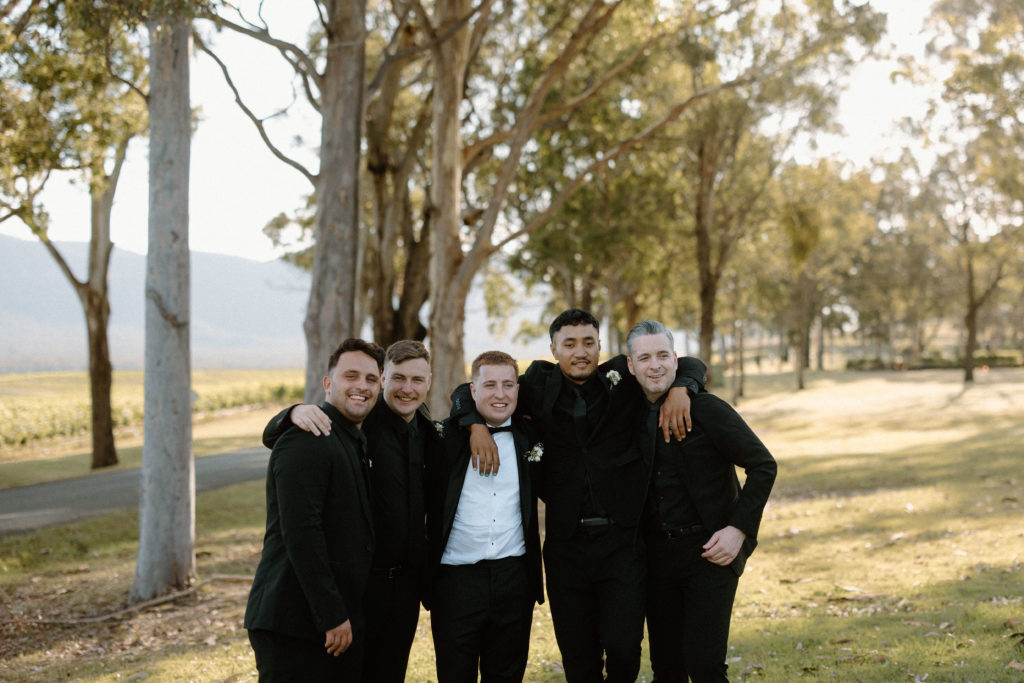 Groom with groomsmen, all wearing black suits,  with arms wrapped around each other smiling