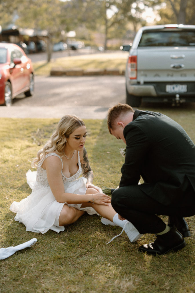 Bride sitting on the grass changing into white sneakers, with the groom helping her